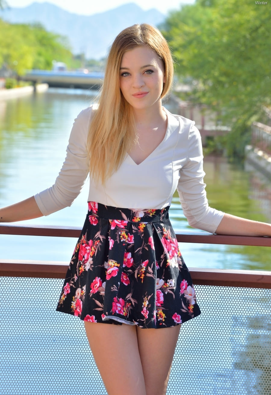 Фото красивой девушки блондинки сдлинным волосом в цветастой мини юбке стоит на мостике у реки в белой кофточке опираясь на перила улыбаясь